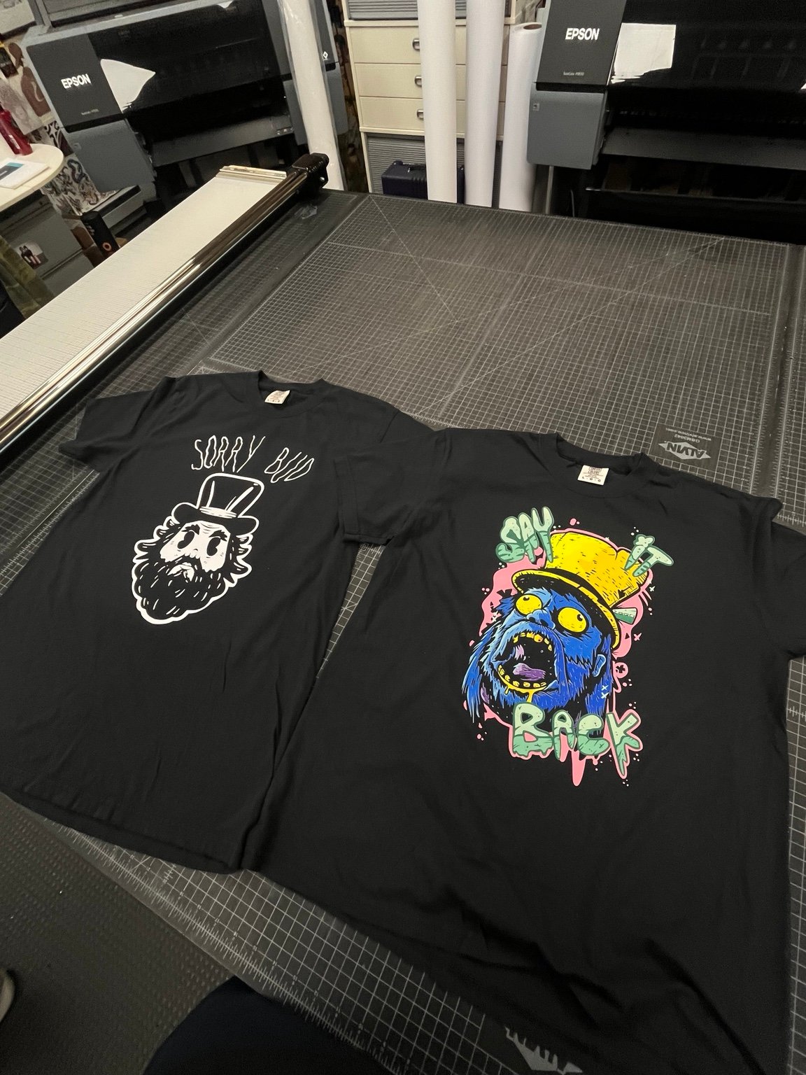 Two custom black graphic shirts on a print shop work table
